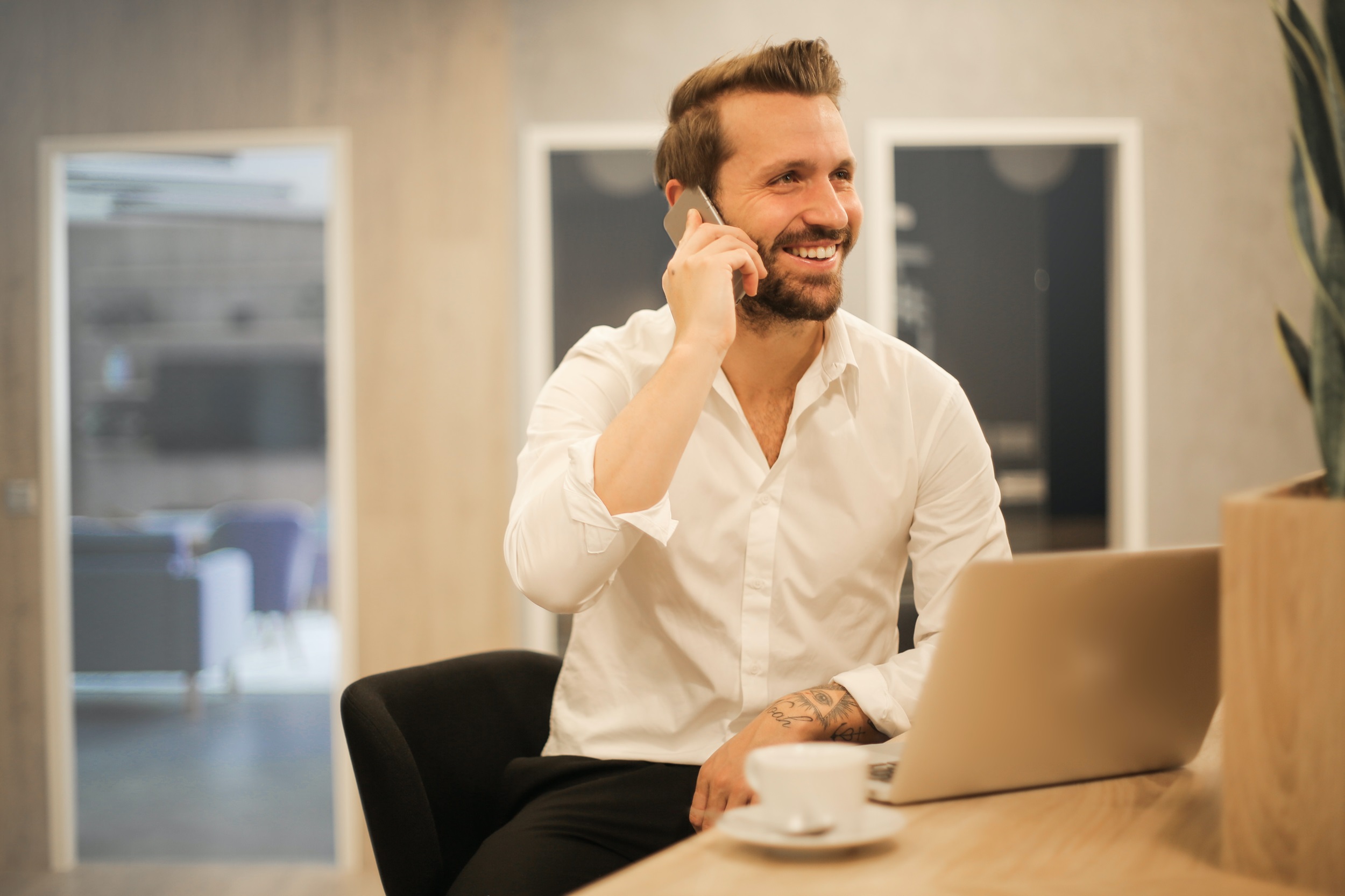 A man in a white button-down shirt smiles while talking on the phone in an office, with a laptop and cup in front of him