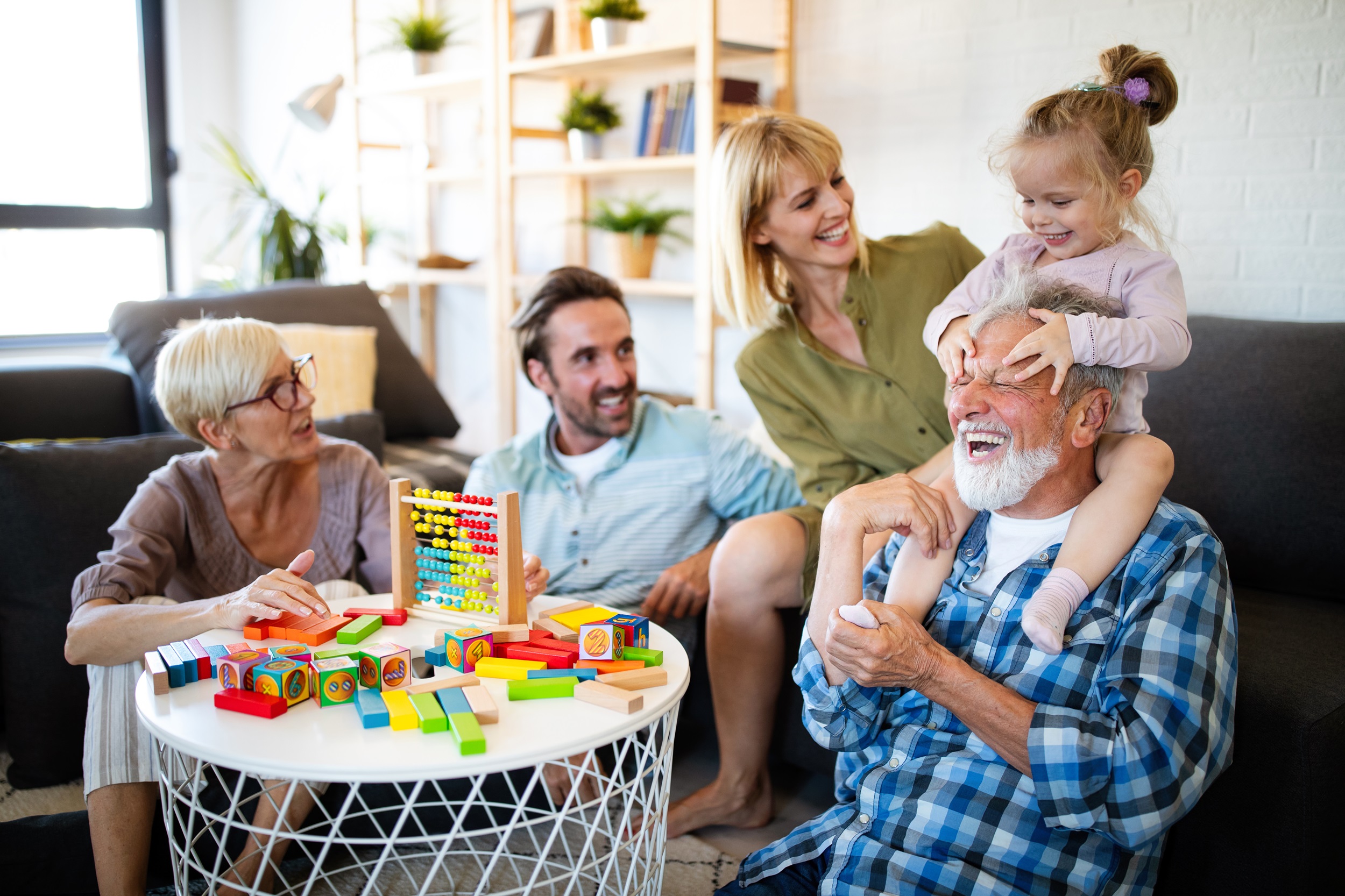 Grandpa holding his granddaughter on his shoulder, with blocks and toys on a central table, and five people laughing together